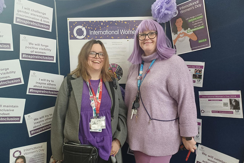 Sam and Rae in front of an International Womens Day display. Both are wearing purple