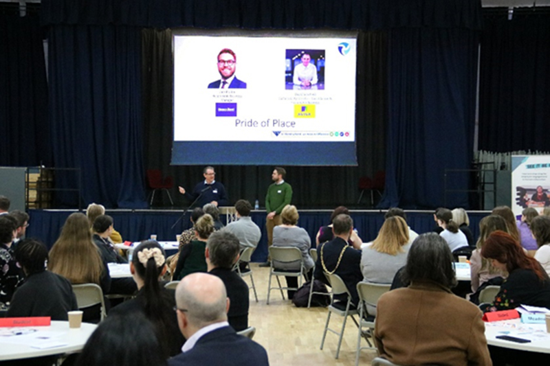 Two people presenting to an audience in front of a large screen in a school hall